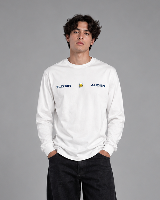 A man stands against a gray studio background wearing a white long-sleeve crewneck sweatshirt with 'FLAVERY' and 'ALDERN' printed on the chest, paired with loose black pants and white sneakers.