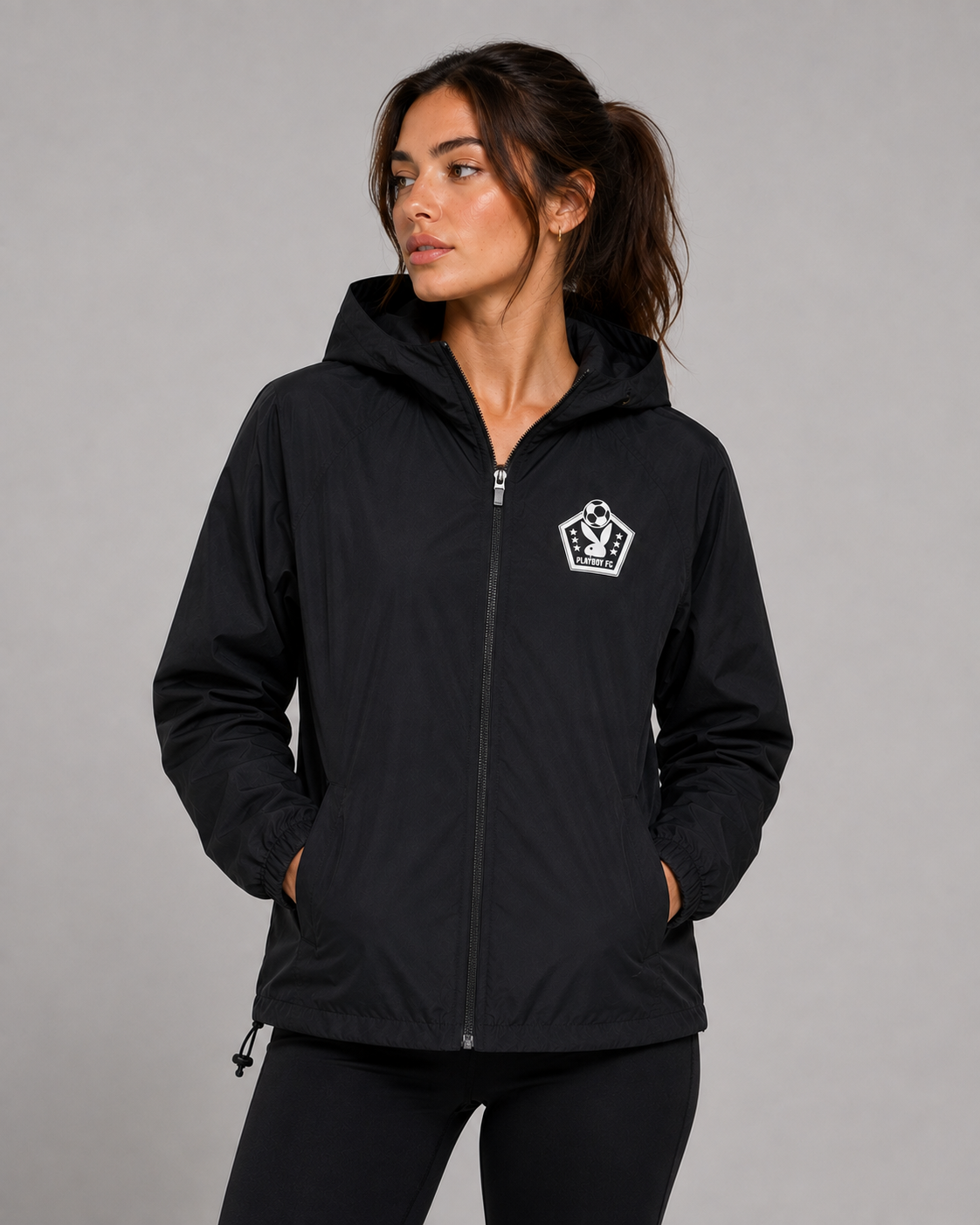 A woman wearing a black hooded zip-up jacket with a white shield logo on the left chest, posed against a solid gray studio background.
