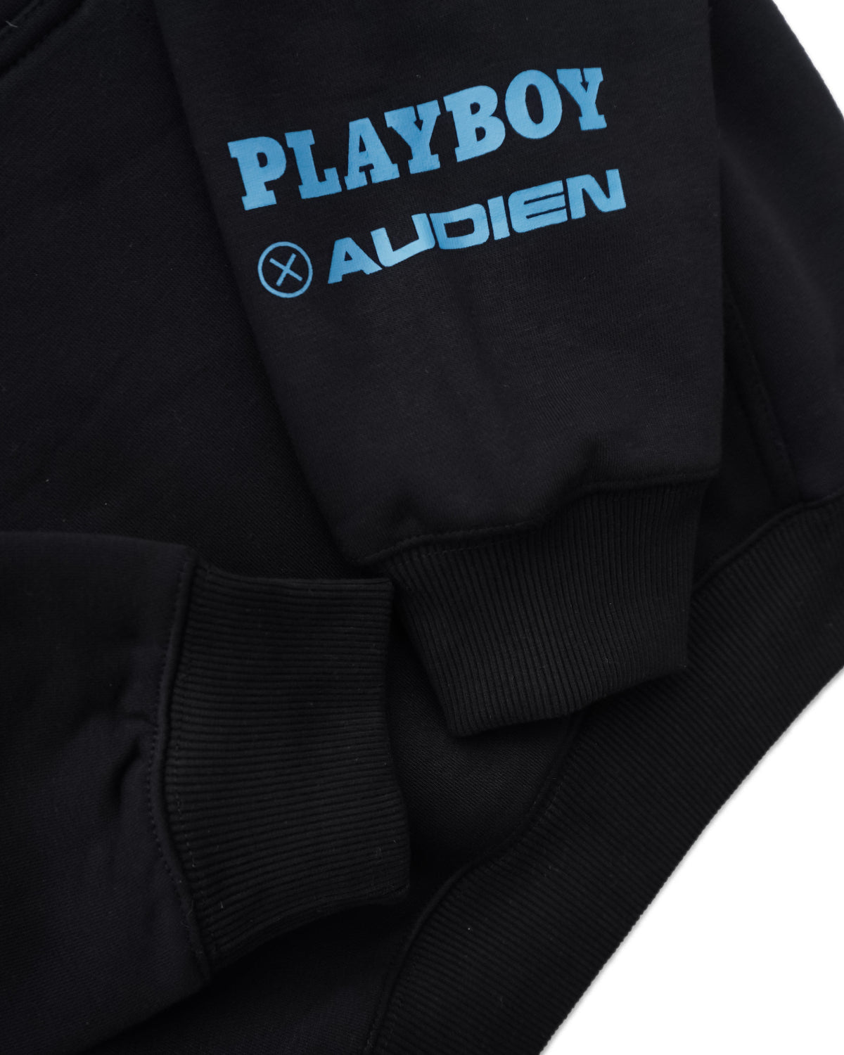 Close-up of a black sweatshirt featuring a light blue graphic print on the sleeve that reads 'PLAYBOY' above 'AUDIEN' with a circular X logo.