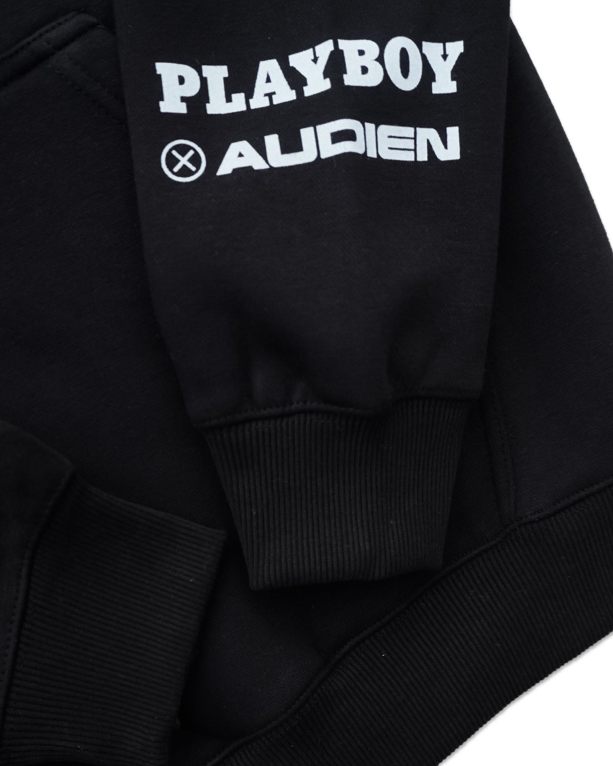 Close-up of a black sweatshirt sleeve featuring a white graphic print that reads 'PLAYBOY' above 'AUDIEN' with a circular cross logo, set against a plain white background.