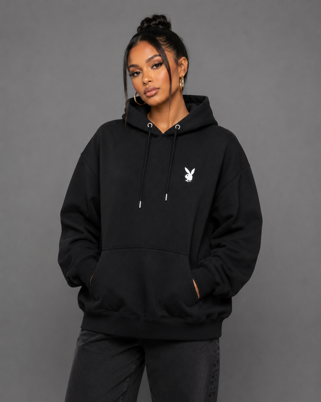 A woman modeling a black pullover hoodie with a small white Playboy bunny logo on the left chest, set against a solid gray studio background.