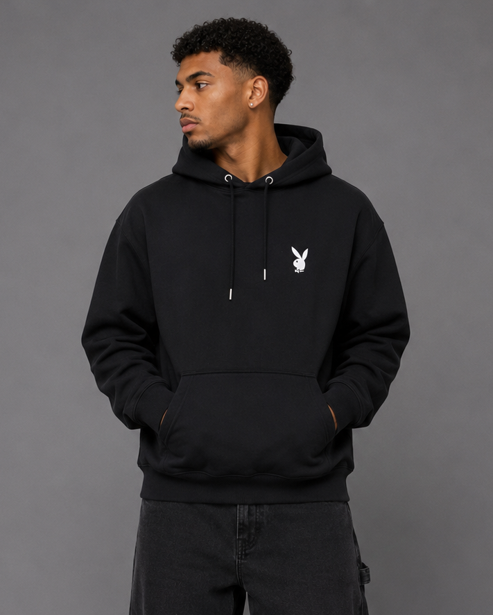 A man wearing a black pullover hoodie with a small white Playboy bunny logo on the left chest, paired with black jeans and white sneakers, standing against a solid gray studio background.