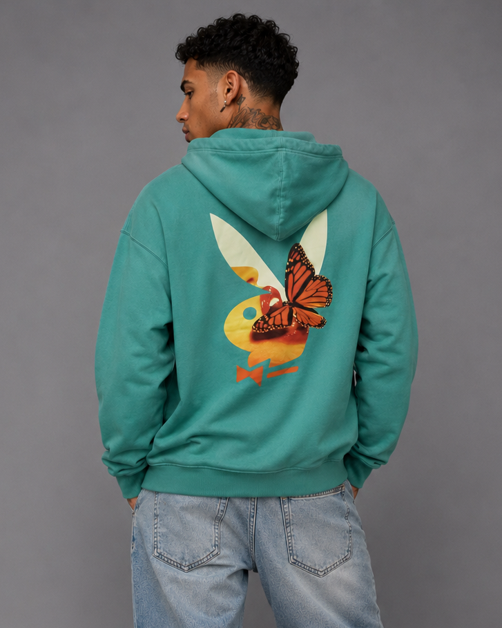 A man wearing a teal green hoodie with a large graphic of a butterfly on a duck on the back, paired with light blue jeans and white sneakers, photographed from behind against a gray studio background.