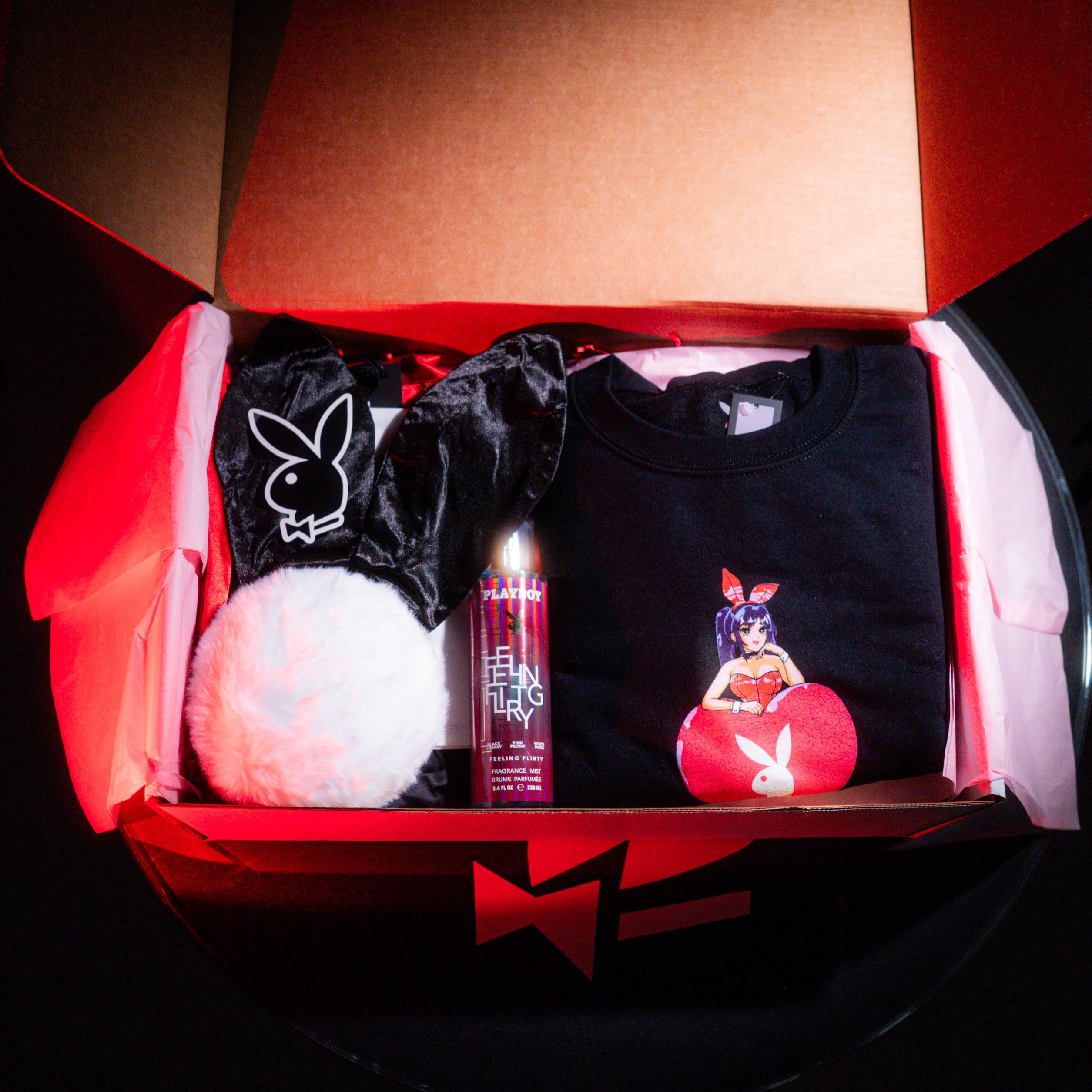 Open box containing a black t-shirt with graphics, a red bottle, and a white plush item on a dark background.