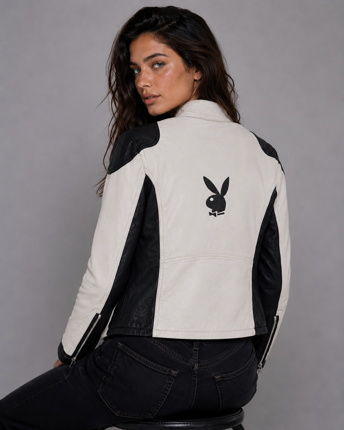 A woman wearing a black and off-white color-blocked jacket with the Playboy bunny logo on the back, photographed against a solid gray studio background. #Women