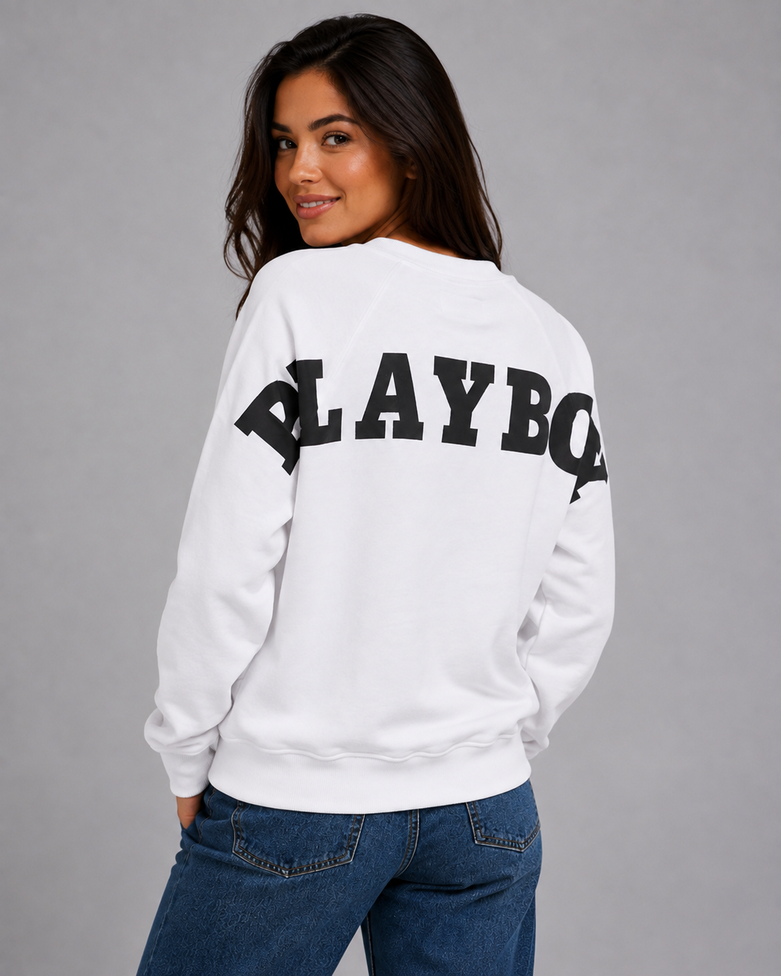 A woman wearing a white sweatshirt with 'PLAYBOY' printed in large black letters across the back, paired with blue jeans, photographed against a plain gray studio background. #Women