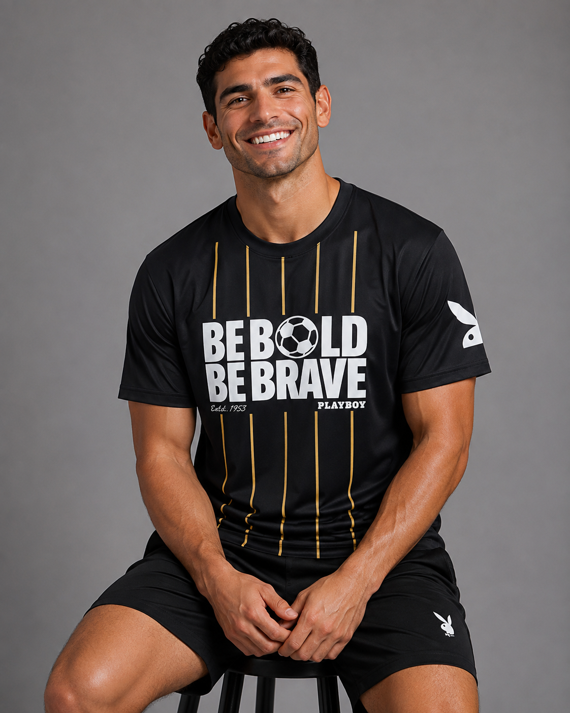 A smiling man seated on a stool wearing a black short-sleeve athletic T-shirt with bold white text reading 'BE BOLD BE BRAVE,' vertical gold stripes, and a white Playboy logo on the sleeve, against a plain gray studio background.