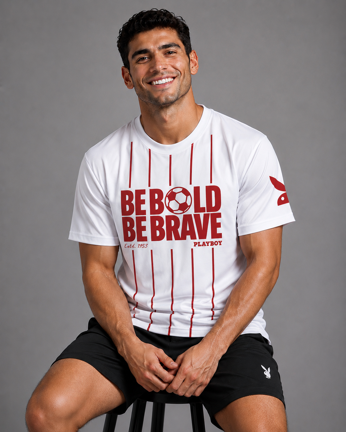 A smiling man seated on a stool wearing a white short-sleeve T-shirt with red vertical stripes and large red text reading 'BE BOLD BE BRAVE' on the front, plus a small Playboy logo on the sleeve and shorts.
