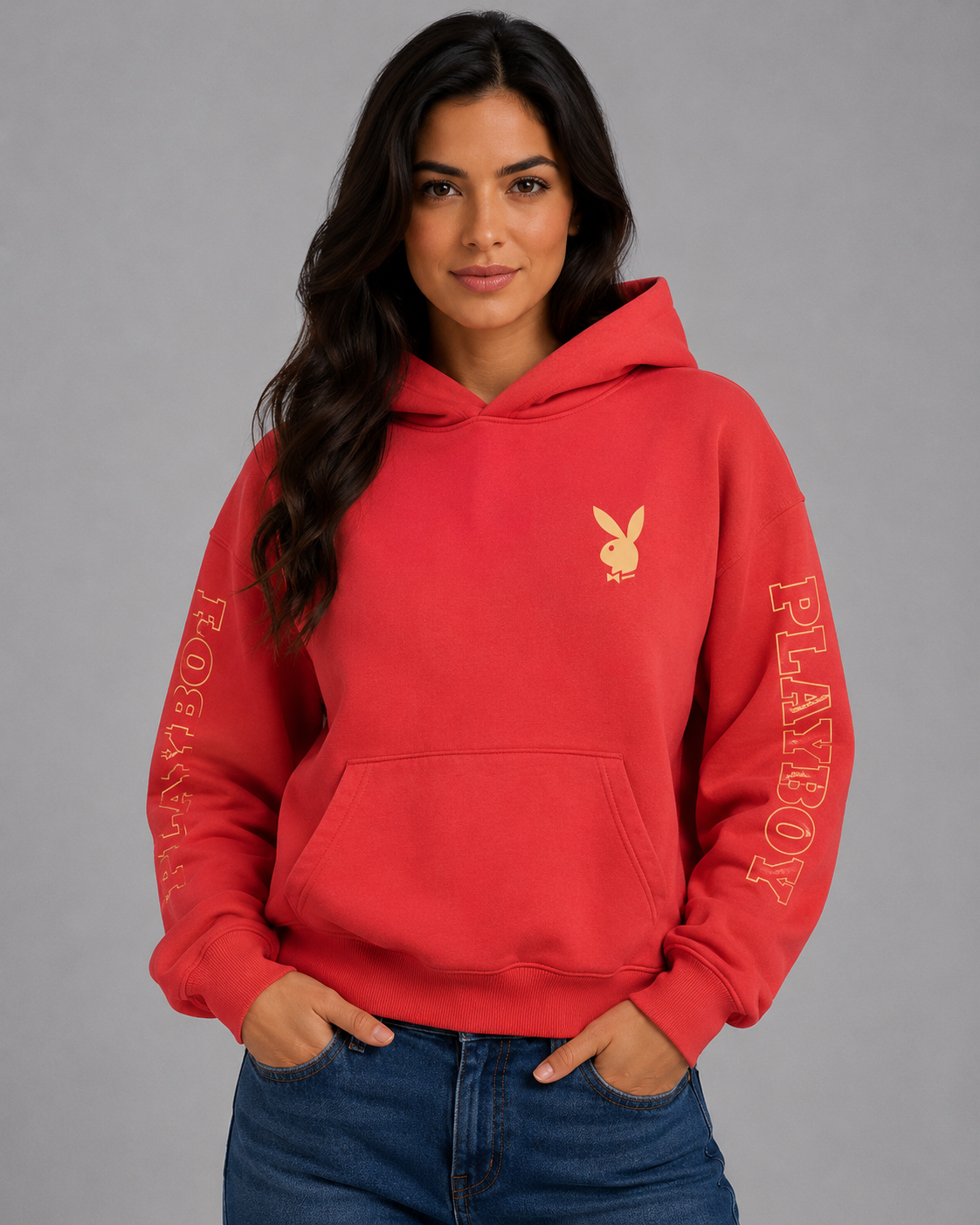 A woman modeling a red pullover hoodie with a gold Playboy bunny logo on the chest and 'PLAYBOY' printed down both sleeves, paired with blue jeans against a solid gray studio background. #Women