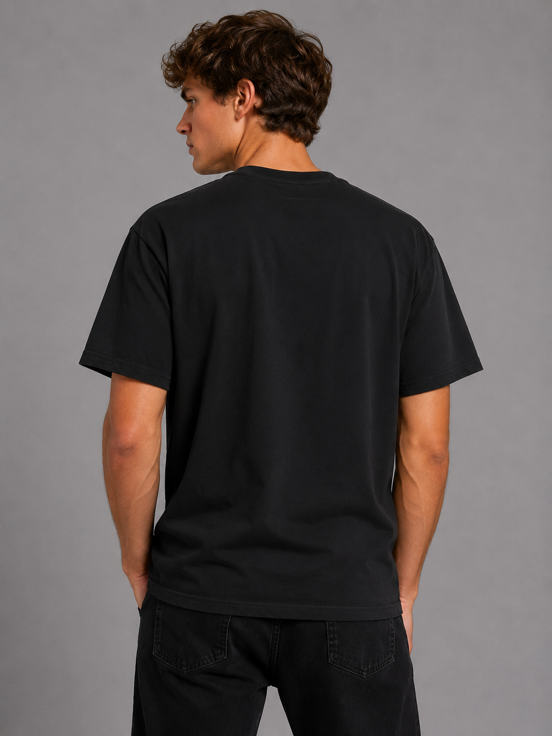 A man viewed from the back wearing a plain black short-sleeve crewneck t-shirt and black pants, standing against a solid gray studio background.