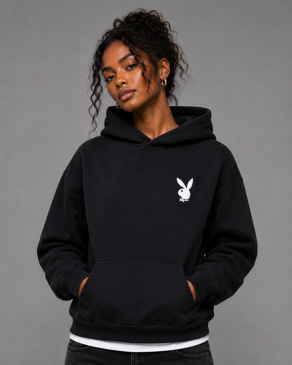 A woman wearing a black pullover hoodie with the white Playboy bunny logo on the left chest, paired with black jeans, against a solid gray studio background. #Women