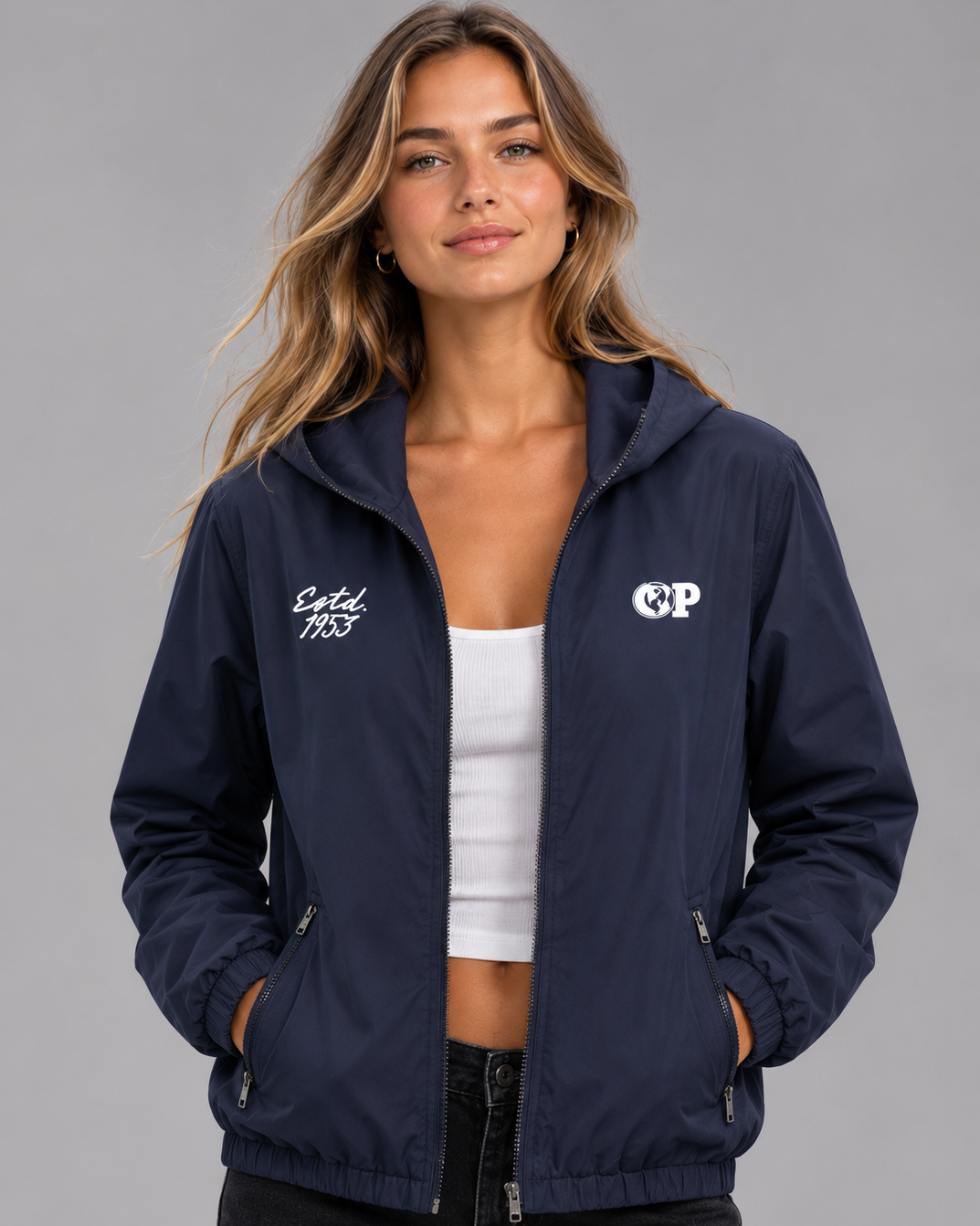 A woman wearing a navy blue zip-up hooded jacket with white embroidered logos on the chest, paired with a white crop top and black pants, photographed against a solid gray studio background. #Women