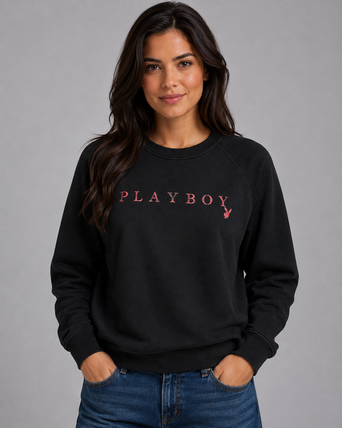A woman wearing a black crewneck sweatshirt with a pink embroidered 'PLAYBOY' logo and rabbit head on the chest, paired with blue jeans, against a solid gray studio background. #Women