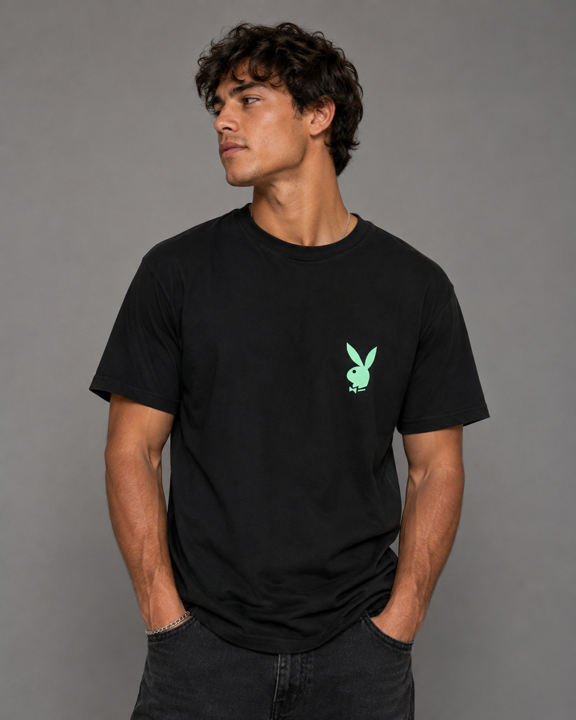 A man wearing a black short-sleeve crewneck t-shirt with a small mint green Playboy bunny logo on the left chest, posed against a solid gray studio background.