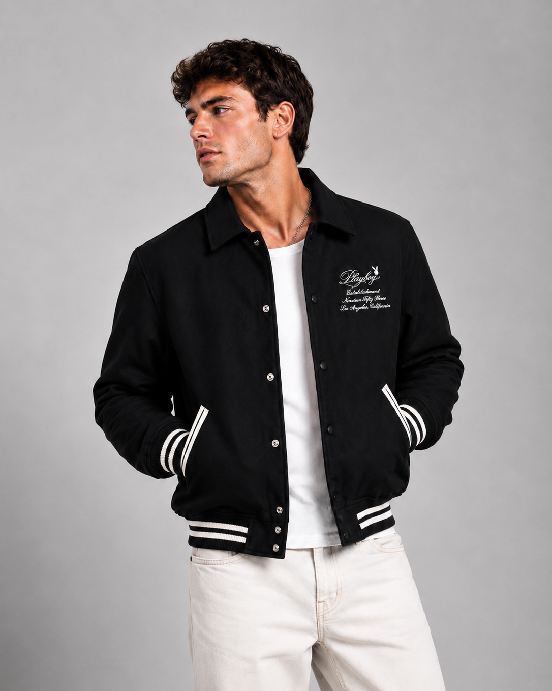 A man wearing a black varsity-style bomber jacket with white striped ribbed cuffs and hem, a white button-up shirt, and off-white trousers, photographed against a neutral gray studio background.