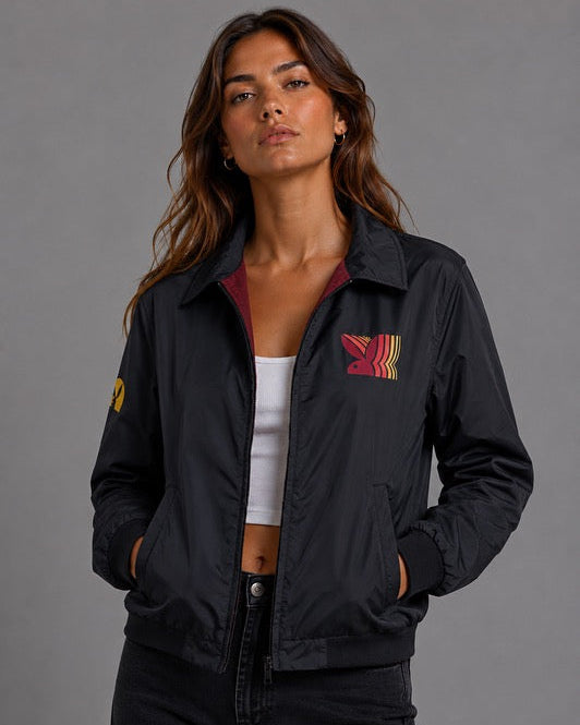 A woman wearing a black bomber jacket with a red and yellow striped wing logo on the left chest, a white crop top, and black jeans, photographed against a solid gray studio background. #Women