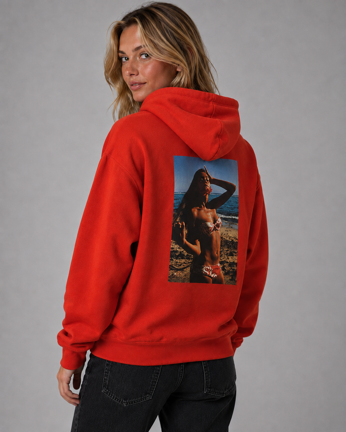 A woman wearing a bright red hoodie from the back, featuring a large rectangular graphic print of a beach scene on the back, paired with black jeans against a solid gray studio background. #Women