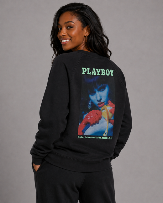 A woman wearing a black sweatshirt with a large back graphic featuring the green 'PLAYBOY' logo and a colorful portrait print, photographed against a plain gray studio background.