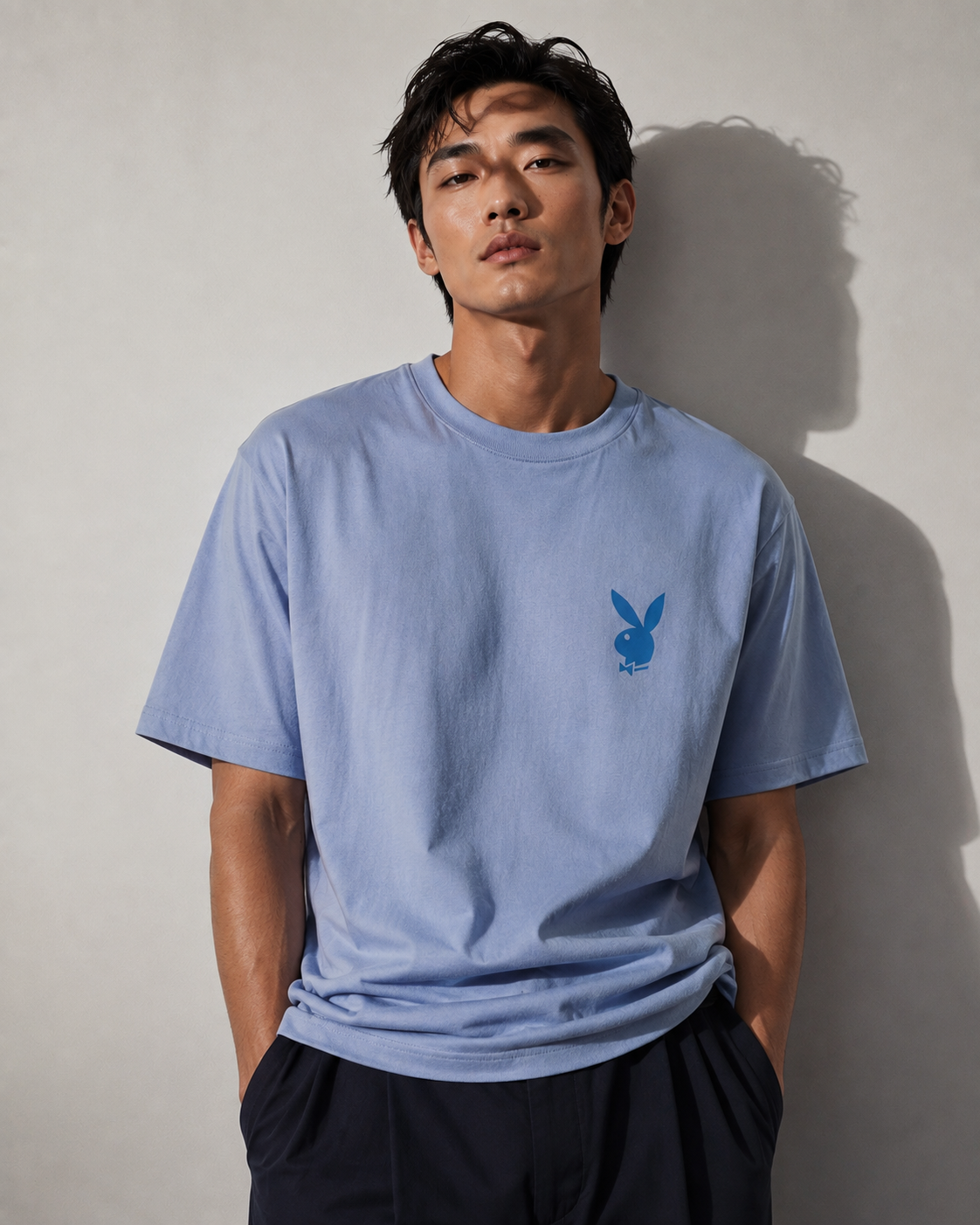 A man wearing a light blue crewneck t-shirt with a small blue Playboy bunny logo on the left chest, paired with dark pants, standing against a neutral gray wall with a soft shadow.