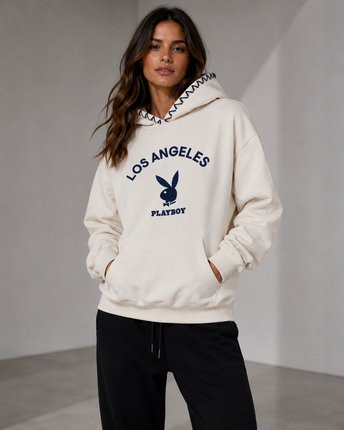 A woman wearing a cream-colored Playboy Los Angeles hoodie with navy blue graphics and black sweatpants, standing against a neutral gray studio background. #Women