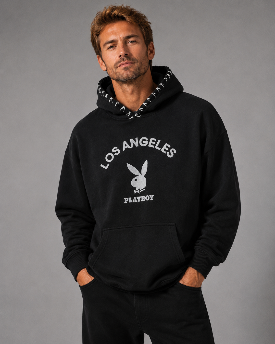 A man wearing a black pullover hoodie with a white Los Angeles Playboy graphic on the front, set against a solid gray studio background.
