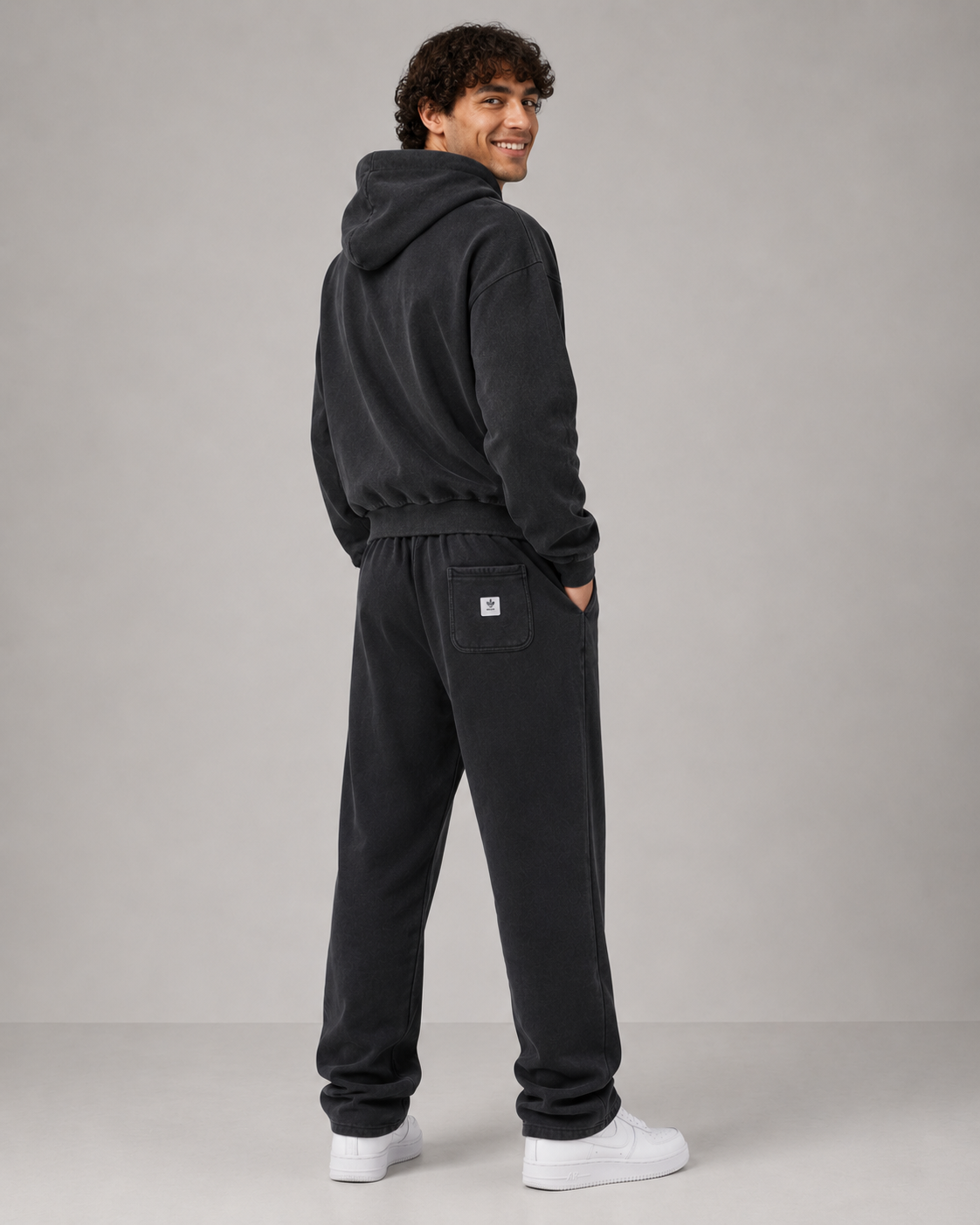 A man wearing a black hooded sweatshirt and matching black sweatpants, shown from the back in a studio setting with a light gray background.