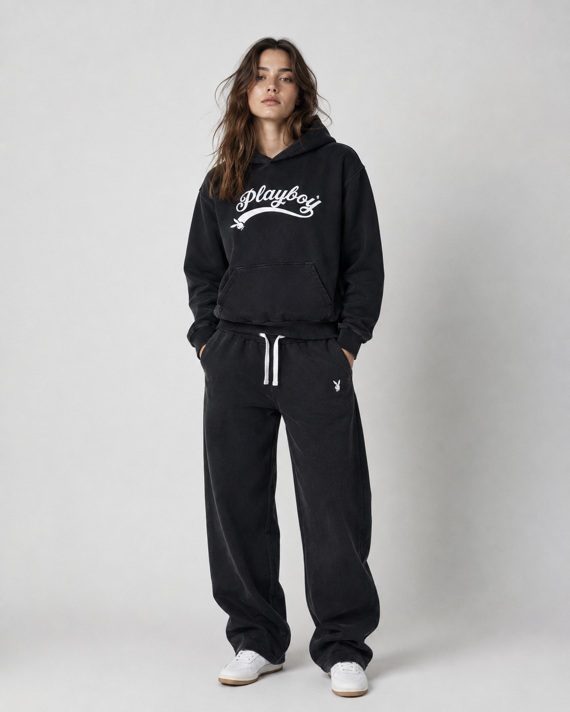 A woman modeling a black Playboy lounge set consisting of a pullover hoodie and matching sweatpants, with white sneakers, against a light gray studio background.