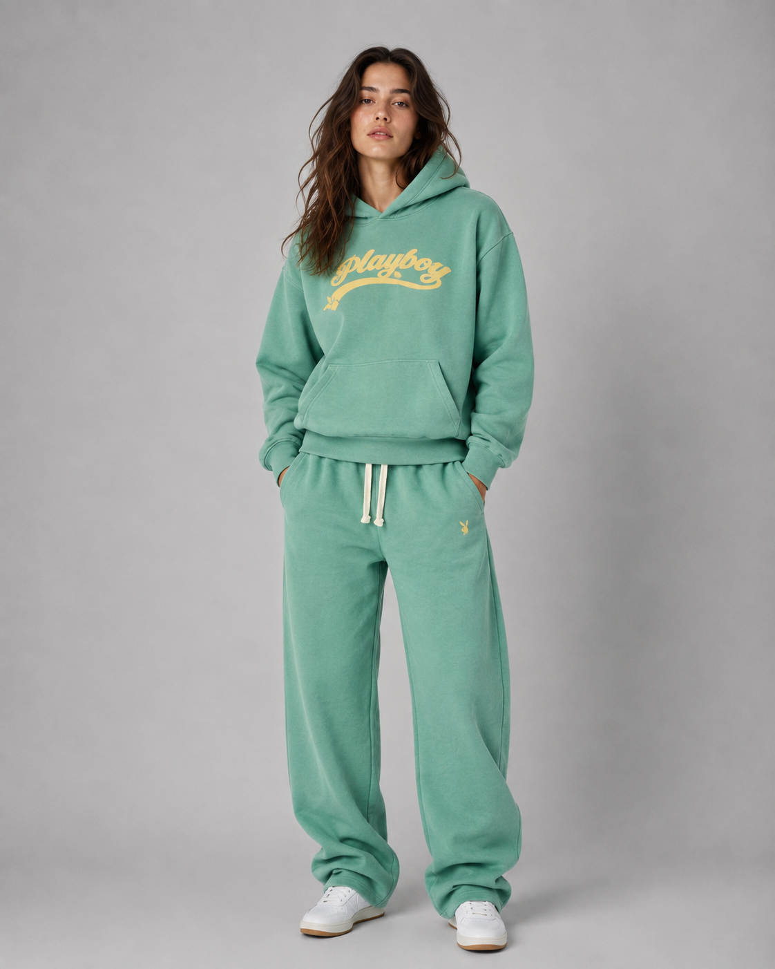 A woman modeling a matching mint green sweatshirt and sweatpants lounge set with white drawstrings and a yellow Playboy logo on the chest, set against a plain gray studio background. #Women