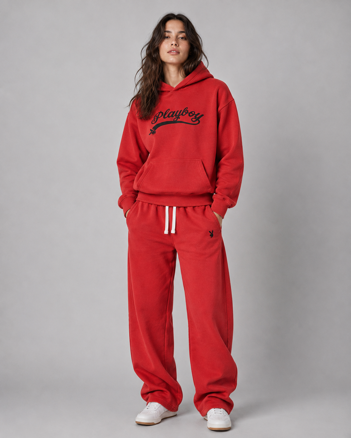 A woman modeling a red sweatshirt and matching sweatpants set from Playboy, featuring a black script logo on the chest and white drawstrings on the pants, set against a plain gray studio background. #Women