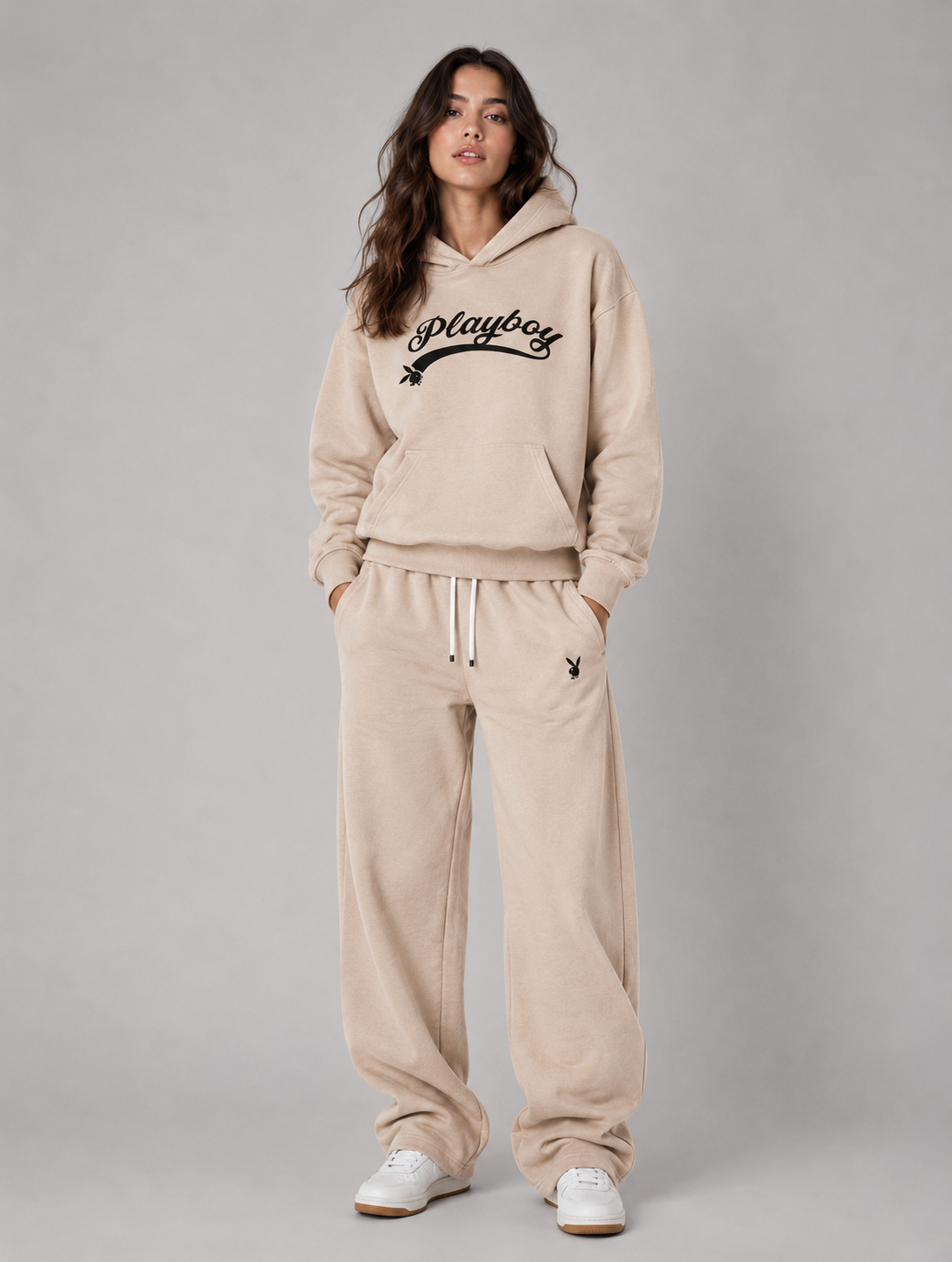 A woman modeling a beige fleece hoodie and matching sweatpants set from Playboy, featuring the brand's logo on the chest and thigh, against a solid gray studio background. #Women