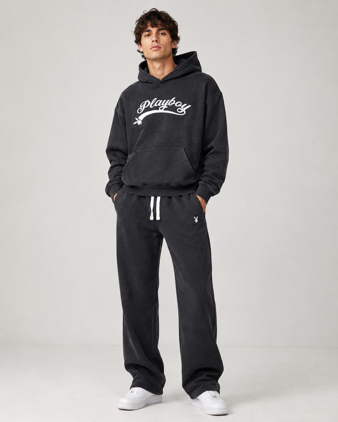 A man modeling a dark charcoal gray hoodie and sweatpants set from Playboy, featuring a white script logo on the chest and white drawstrings, paired with white sneakers against a light gray studio background.