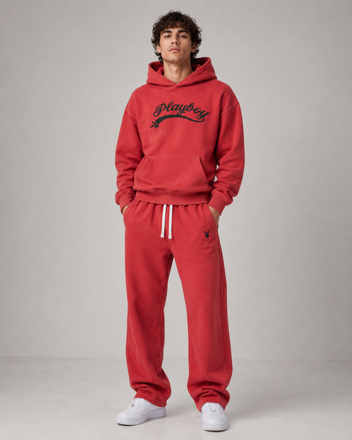 A man modeling a red sweatshirt and matching sweatpants set from Playboy, featuring a black script logo on the chest and white drawstrings on the pants, set against a plain gray studio background.