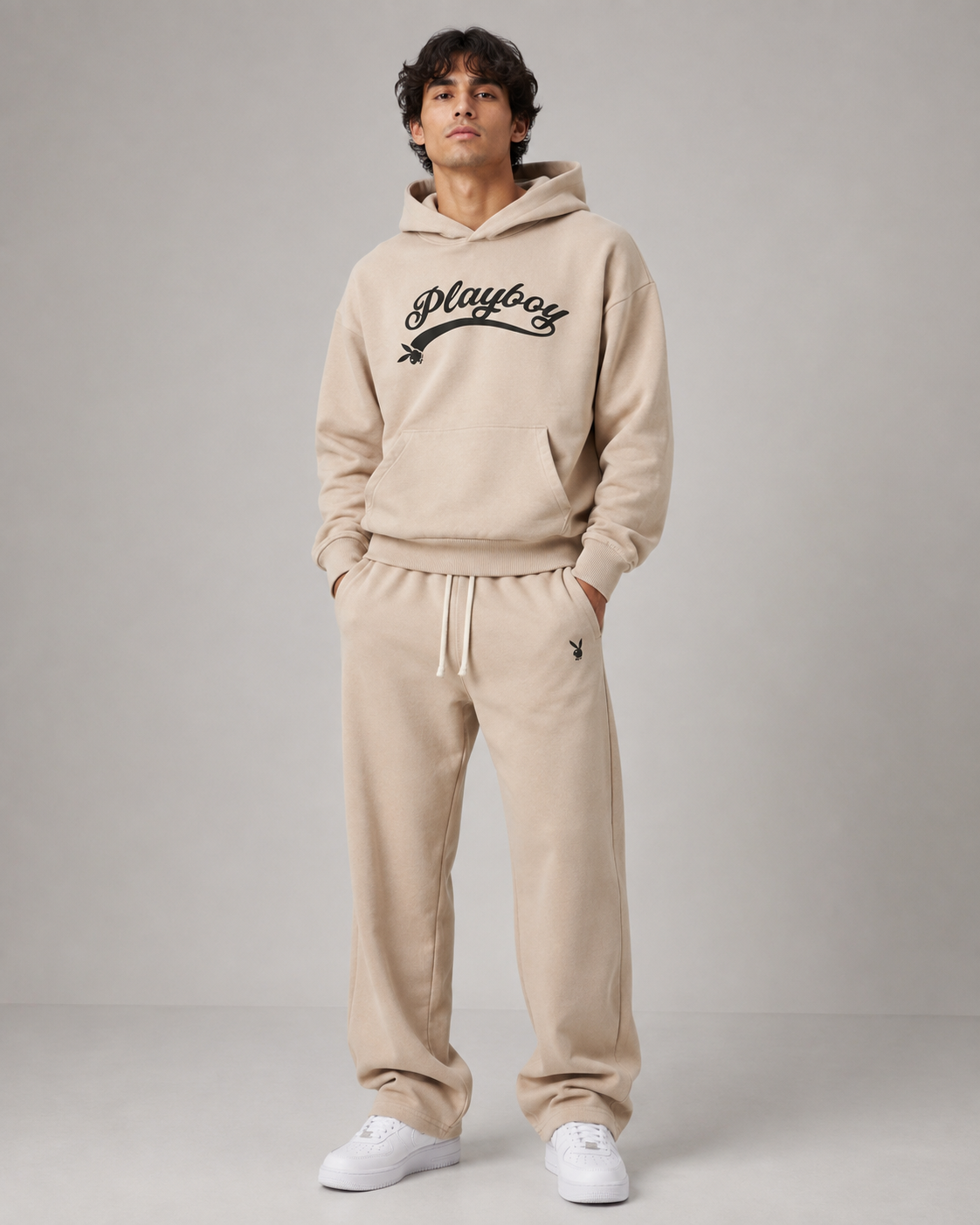 A man modeling a beige two-piece loungewear set consisting of a pullover hoodie with a black 'Playboy' script logo on the chest and matching jogger sweatpants with a drawstring waist, paired with white sneakers against a neutral gray studio background.