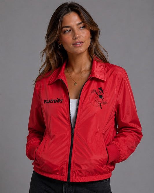 A woman wearing a bright red zip-up jacket with black Playboy branding and graphics, paired with a white top and black jeans, photographed against a solid gray studio background. #Women