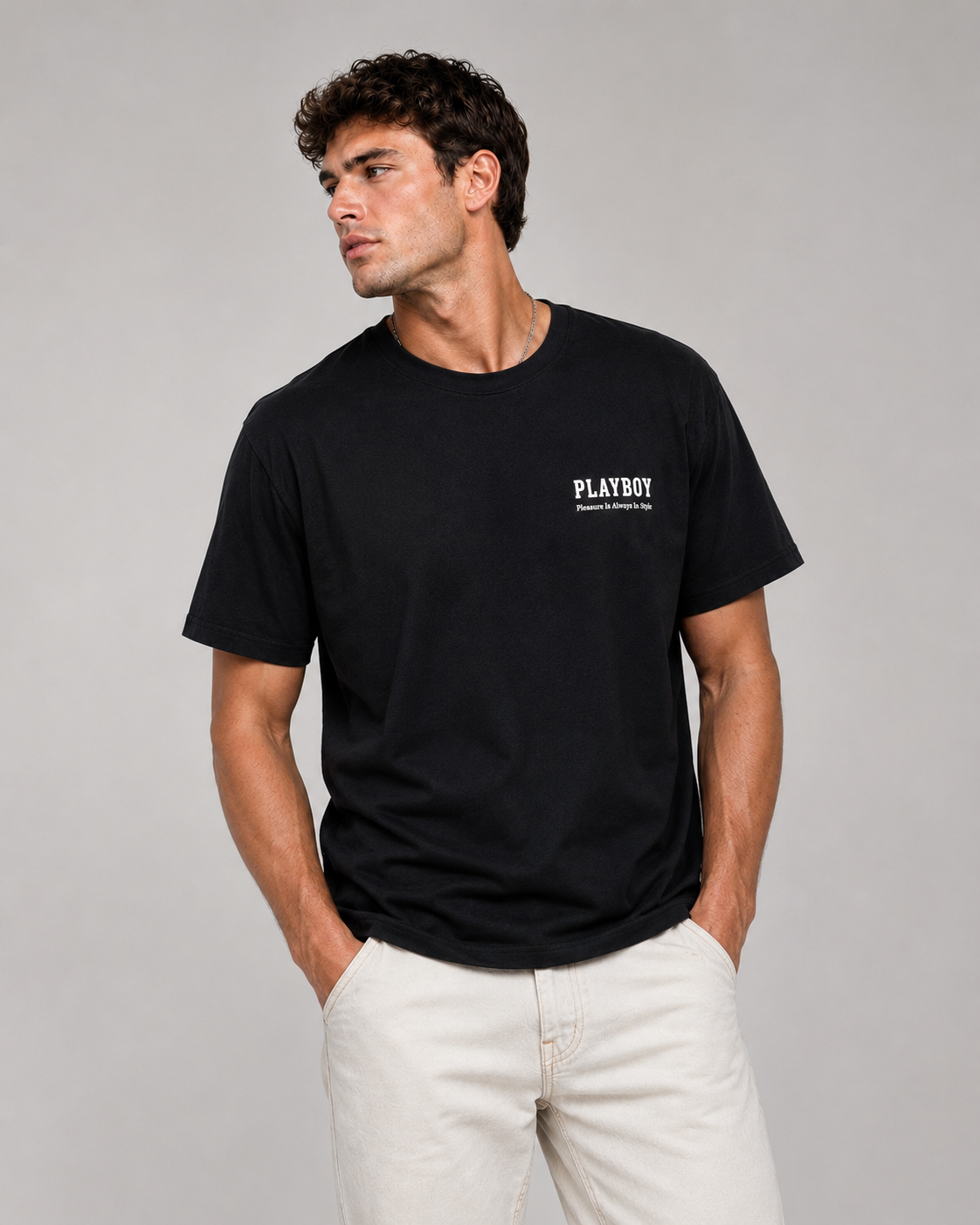 A man wearing a black short-sleeve crewneck t-shirt with a small white PLAYBOY logo on the left chest, paired with off-white trousers, against a solid gray studio background.
