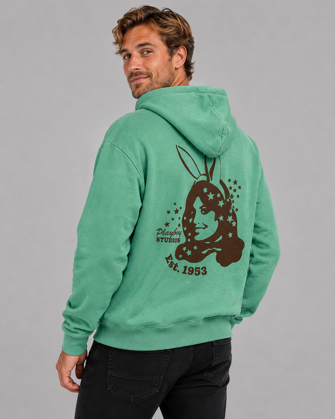 A man wearing a mint green hoodie with a large brown bunny graphic and text on the back, shown from a three-quarters back view against a solid gray studio background.
