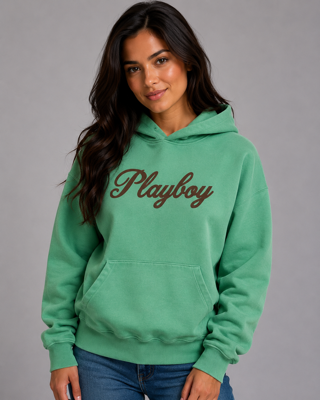 A woman wearing a mint green hoodie with the word 'Playboy' printed across the chest in brown cursive, paired with blue jeans, against a solid gray studio background. #Women