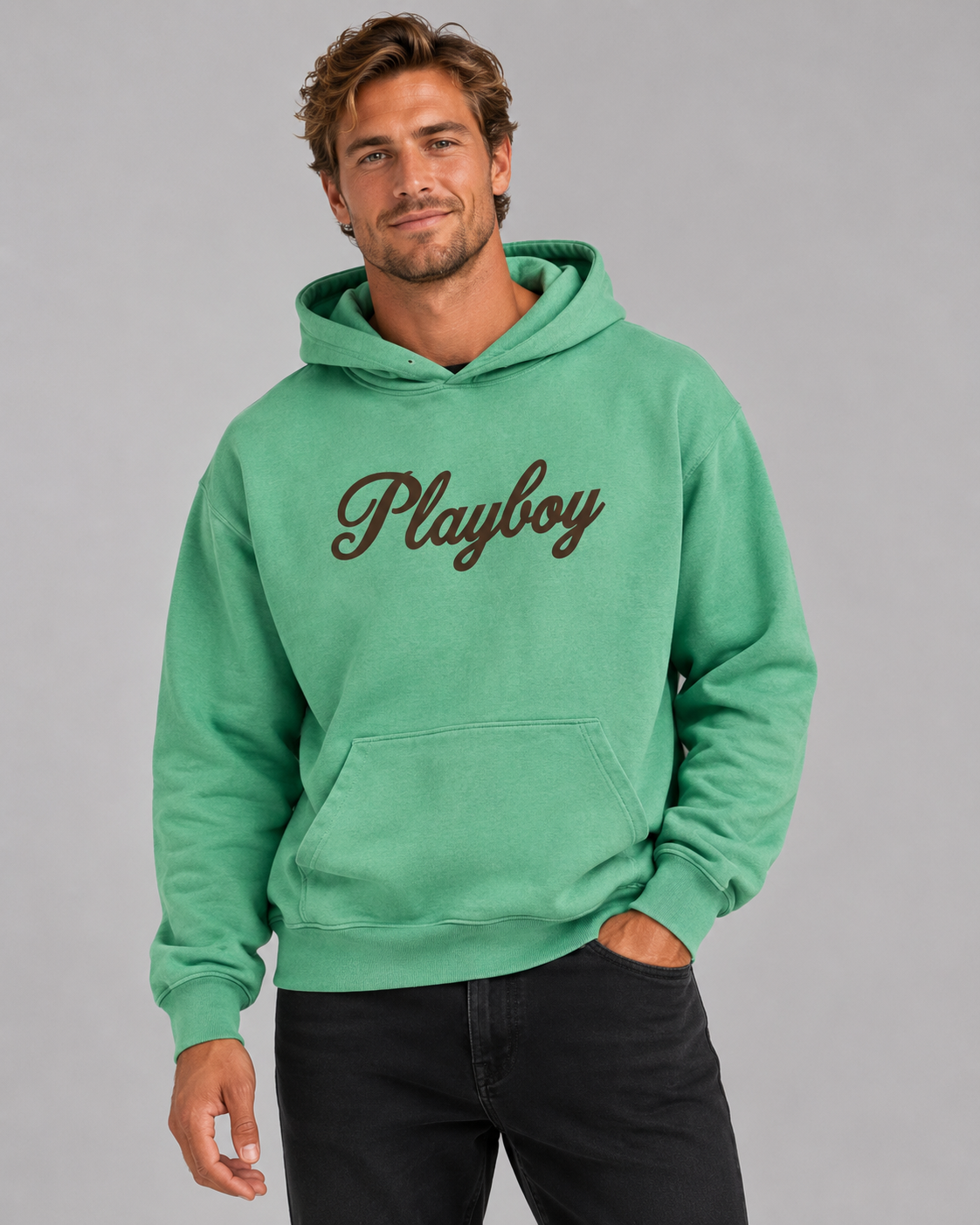 A man wearing a green pullover hoodie with the word 'Playboy' embroidered in dark brown script across the chest, paired with black pants, against a solid gray studio background.