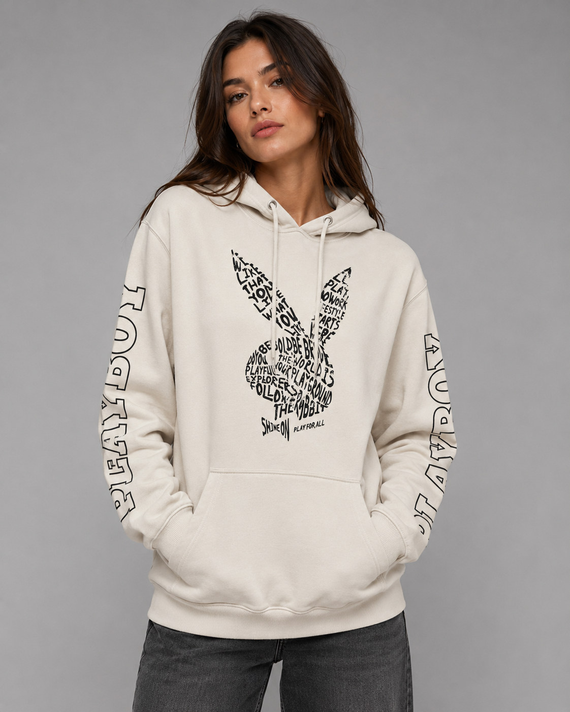 A woman wearing a cream-colored Playboy hoodie with a black graphic print of the brand's bunny logo and 'PLAYBOY' text on the sleeves, paired with dark gray jeans, against a solid gray studio background. #Women