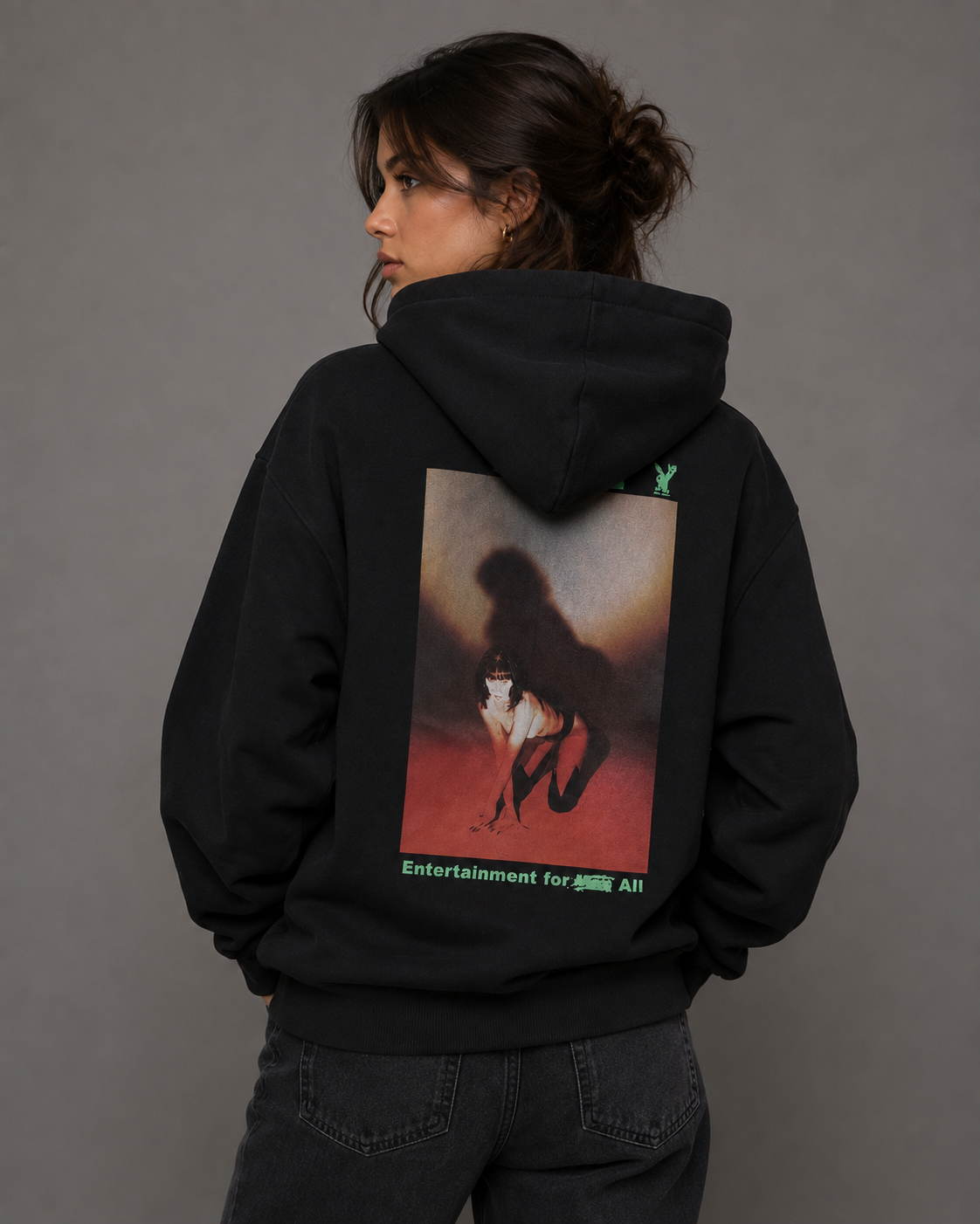 A model shown from the back wearing a black hoodie with a large graphic print on the back and a small green logo on the left shoulder, paired with dark jeans against a solid gray studio background. #Women