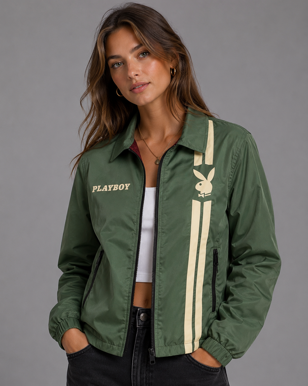 A woman wearing an olive green bomber jacket with cream-colored stripes and the Playboy bunny logo on the chest, paired with a white crop top and black jeans, against a solid gray studio background. #Women