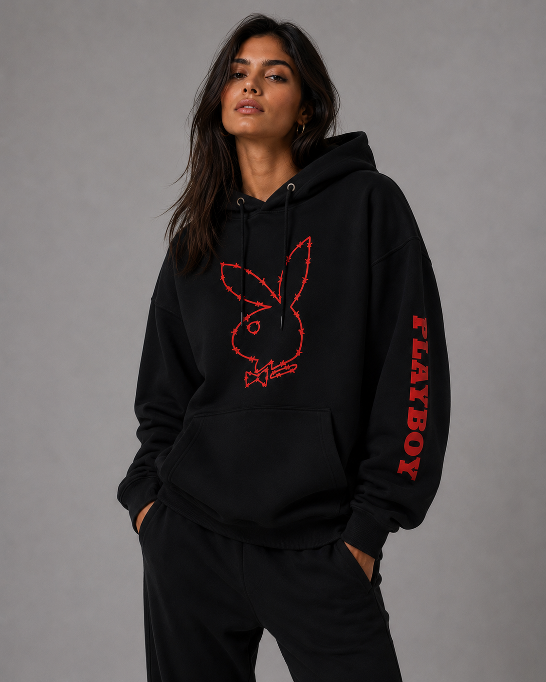 A woman wearing a black pullover hoodie with a red Playboy bunny logo on the chest and 'PLAYBOY' text down one sleeve, paired with black sweatpants, against a plain gray studio background.
