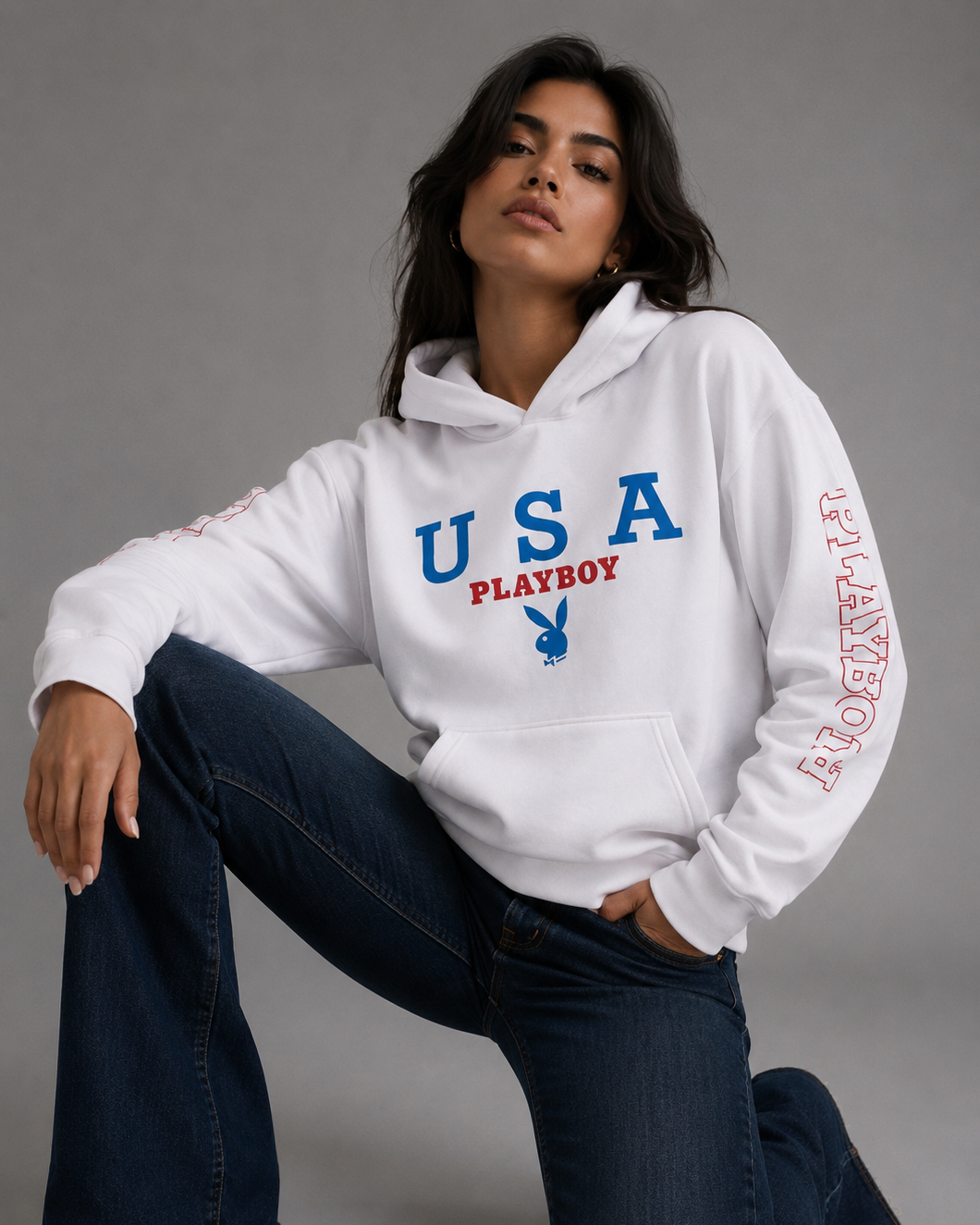 A woman kneels on a gray studio floor wearing a white hoodie with 'USA PLAYBOY' printed in blue and red on the front, plus red text down one sleeve. She is also wearing dark blue flared jeans. #Women