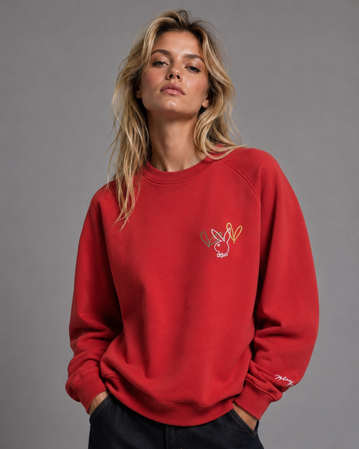 A woman wearing a bright red crewneck sweatshirt with a small embroidered bunny graphic on the left chest, paired with dark jeans, against a solid gray studio background.