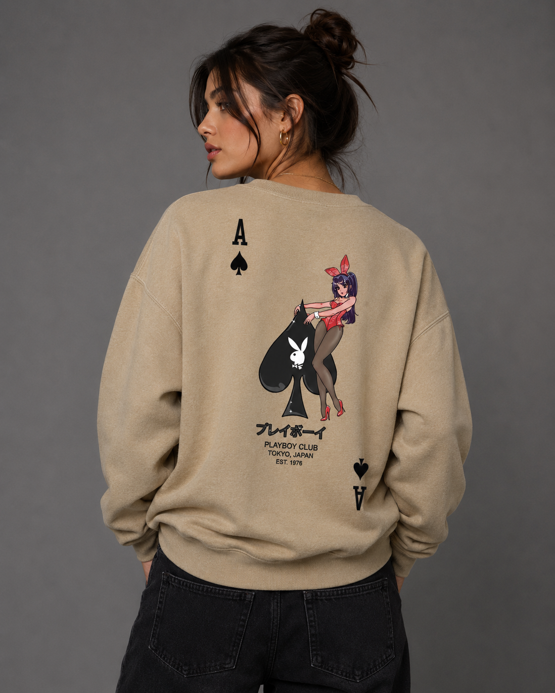 A woman shown from the back wearing a beige sweatshirt with a large Playboy back graphic featuring an Ace of Spades and a cartoon character, paired with dark jeans against a solid gray studio background. #Women