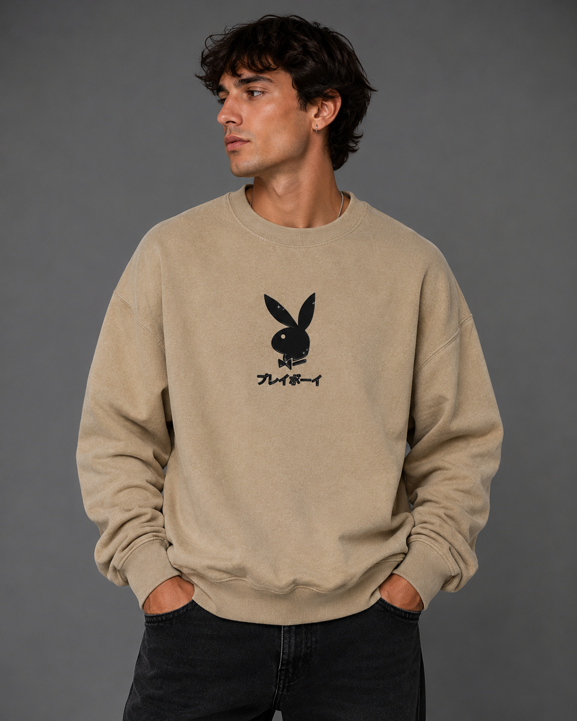 A man wearing a beige crewneck sweatshirt with a black Playboy bunny logo and Japanese text on the chest, paired with dark jeans, against a solid gray studio background.