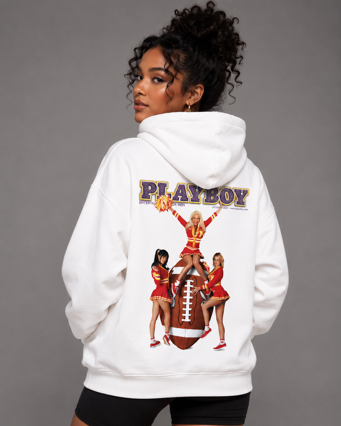 A woman modeling a white Playboy hoodie from the back, featuring a large graphic print of cheerleaders and a football with the 'PLAYBOY' logo. She is wearing black athletic shorts against a solid gray studio background.