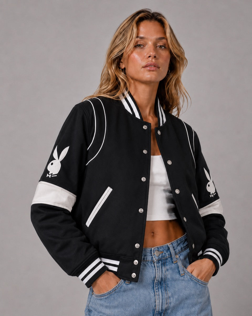 A woman modeling a black varsity-style bomber jacket with white stripes and white Playboy bunny logos on the sleeves, paired with a white crop top and light blue jeans against a gray studio background.