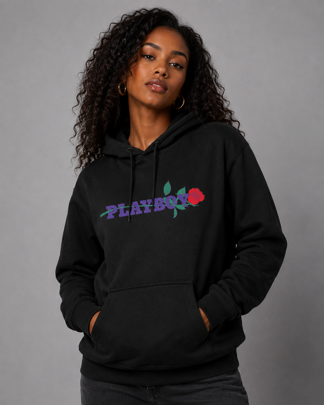 A woman with curly hair wearing a black pullover hoodie with a colorful 'PLAYBOY' logo and red rose graphic on the chest, posed against a solid gray studio background.