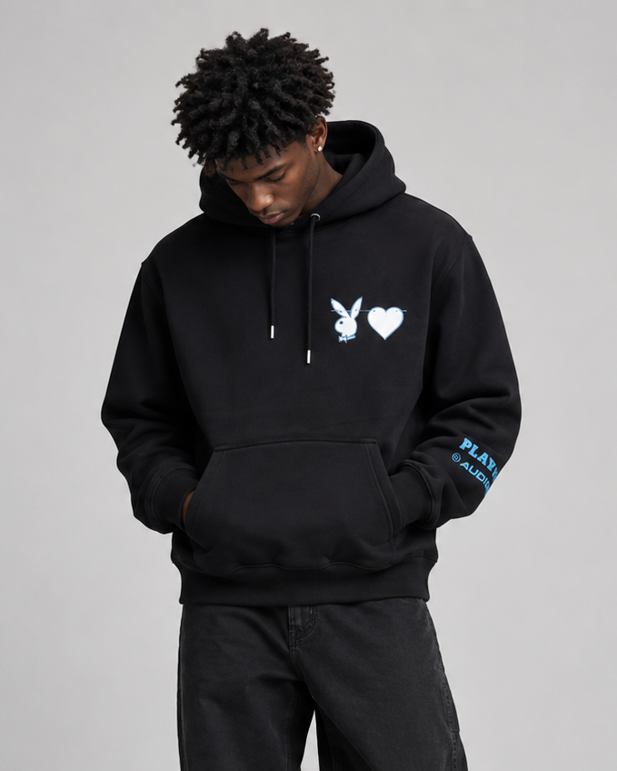 A man modeling a black hoodie and matching black sweatpants against a light gray studio background. The hoodie features a white bunny and heart logo on the chest and blue text on the sleeve.